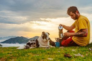 young mountaineer man has breakfast drinking coffee with his dog in the mountains